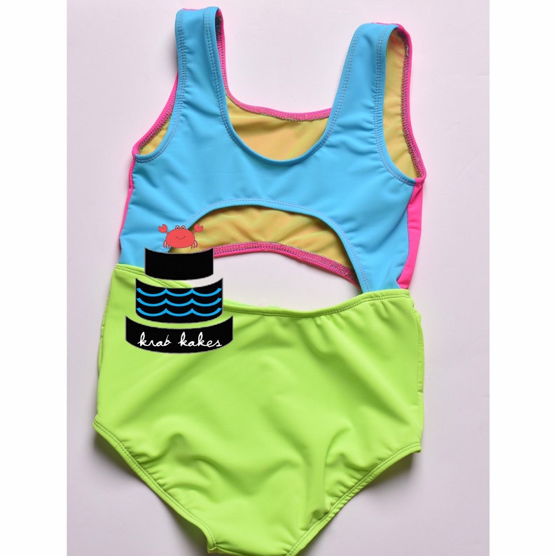 May include: A bright green swimsuit with a blue and pink top. The top has a cutout in the back and a crab cake design on the front. The text "krab kakes" is printed on the swimsuit.