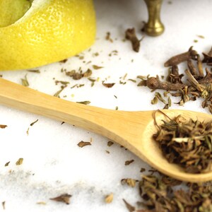 May include: A wooden spoon filled with dried herbs and spices, with additional herbs and cloves scattered on a white surface.
