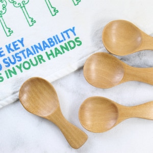 May include: Four small wooden spoons with rounded bowls and short handles. The spoons are arranged on a white surface with a white towel with the text "The key to sustainability is in your hands" printed in green and blue.