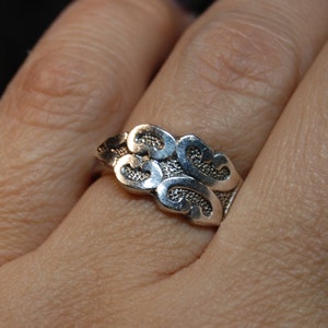 May include: A silver ring with a swirling, ornate design. The ring is worn on a person's finger.