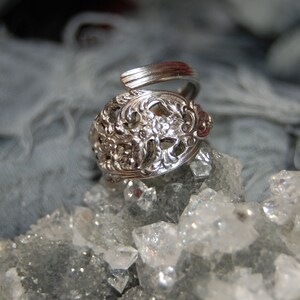 Pode incluir: Um anel de prata com um delicado design floral, descansando sobre um leito de cristais brancos brilhantes.