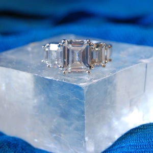 May include: A silver ring with three large, clear, emerald-cut gemstones set in a row. The ring is resting on a clear, crystalline surface.