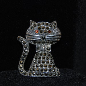 以下が含まれることがあります： 黒と赤のラインストーンが付いた銀製の猫のブローチ。猫は蝶ネクタイをしています。