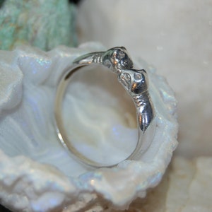 May include: A silver ring with two intertwined animal heads, possibly snakes, facing each other. The ring is set against a white background.