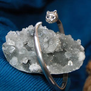 May include: A silver bracelet with a cat head charm. The bracelet is on a bed of white crystals.