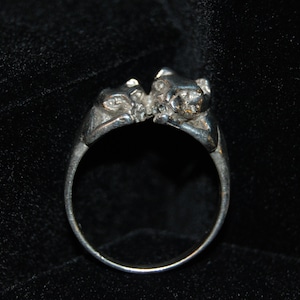 May include: A silver ring with two animal figures, possibly cats, facing each other. The ring is made of a smooth, polished metal.