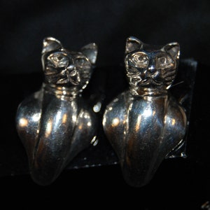 May include: A pair of silver-coloured cat-shaped earrings. Each earring features a detailed cat head with pointed ears and a stylized body. The earrings are set against a dark background, highlighting their metallic sheen.