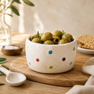 May include: A round, off-white ceramic bowl featuring small, colourful polka dots in shades of blue, red, green, and purple. The bowl is filled with green olives, some garnished with small sprigs of fresh rosemary, creating a simple food presentation.