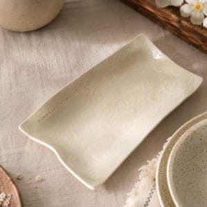 Puede incluir: Una bandeja rectangular de cerámica color blanco hueso con un sutil patrón floral en relieve en la superficie. El texto "HANDMADE WITH LOVE" está estampado en el lado izquierdo de la bandeja, que presenta bordes ligeramente curvados y un acabado brillante.