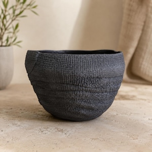 Peut inclure: Un bol gris anthracite foncé avec une surface texturée et irrégulière présentant des crêtes horizontales et une finition mate. Le bol possède une large ouverture et une base arrondie, offrant un aspect organique et tactile d'une couleur uniforme.
