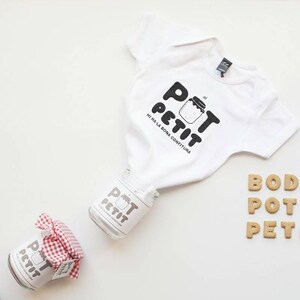 Puede incluir: Un body blanco para bebé con texto negro que dice "Pot Petit" y una pequeña ilustración de un tarro. El body está rodeado de pequeñas galletas que deletrean "Body Pot Petit".
