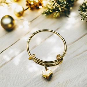 May include: A gold bangle bracelet with a heart charm. The bracelet is on a white wooden surface.