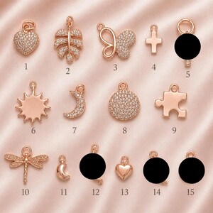 May include: A collection of fifteen rose gold-toned metal charms featuring various shapes including a heart, leaf, butterfly, cross, sun, crescent moon, circle, puzzle piece, and dragonfly, some encrusted with small clear stones and numbered 1 to 15.