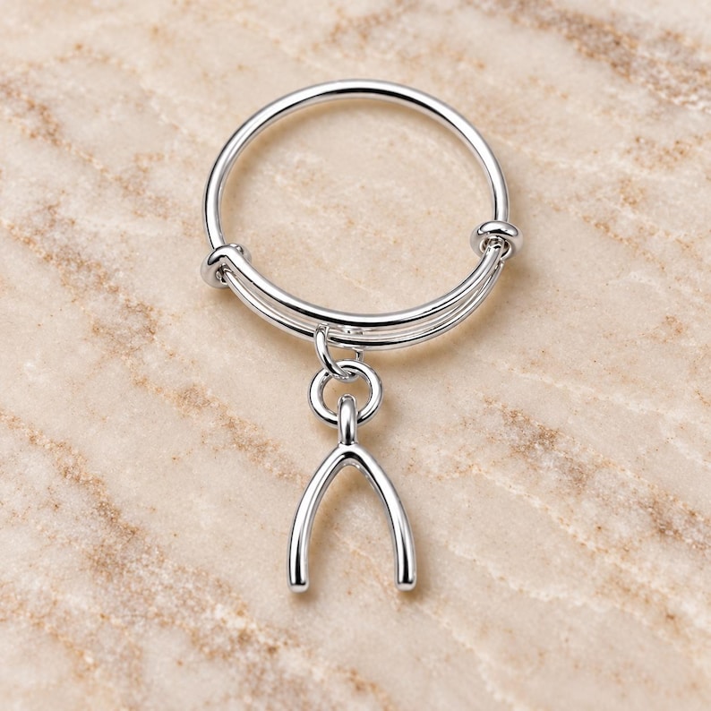 May include: A silver-toned adjustable ring featuring a smooth, polished finish. The band is composed of two thin, overlapping wires that slide to adjust the size. A small, Y-shaped wishbone charm hangs from a circular jump ring attached to the band.