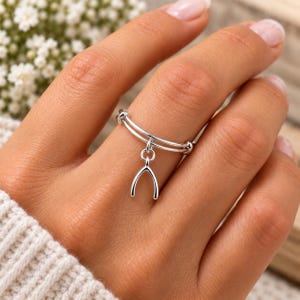 May include: A silver-toned ring featuring a double-band design and a dangling wishbone charm. The ring is constructed from a polished metallic material and displays a simple, clean aesthetic with a singular charm suspended from the center.