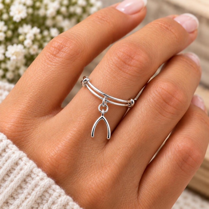 May include: A silver-toned ring featuring a double-band design and a dangling wishbone charm. The ring is constructed from a polished metallic material and displays a simple, clean aesthetic with a singular charm suspended from the center.