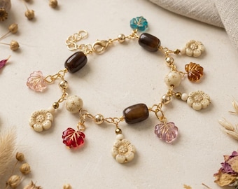 May include: A gold-tone chain charm bracelet featuring dark brown cylindrical beads, small gold-tone spheres, and various dangling charms including cream-colored carved flowers, translucent red, pink, blue, and amber-colored leaves.