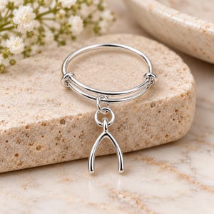 May include: A silver-toned adjustable ring featuring a thin, circular band with a dangling wishbone charm. The metal has a polished, reflective finish, and the design includes a small jump ring connecting the charm to the main band structure.