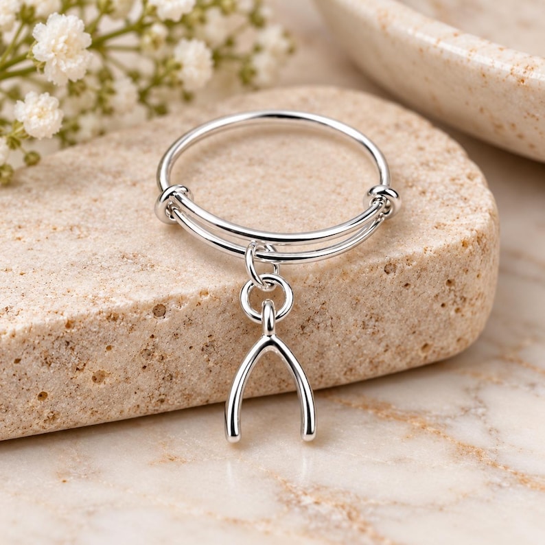 May include: A silver-toned adjustable ring featuring a thin, circular band with a dangling wishbone charm. The metal has a polished, reflective finish, and the design includes a small jump ring connecting the charm to the main band structure.