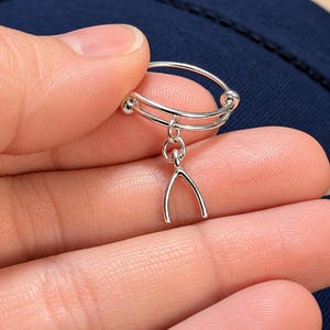 May include: A silver-toned adjustable ring featuring a delicate wishbone charm dangling from a small circular link. The band consists of two thin, parallel silver wires that overlap and terminate in small, smooth spherical beads at each end.
