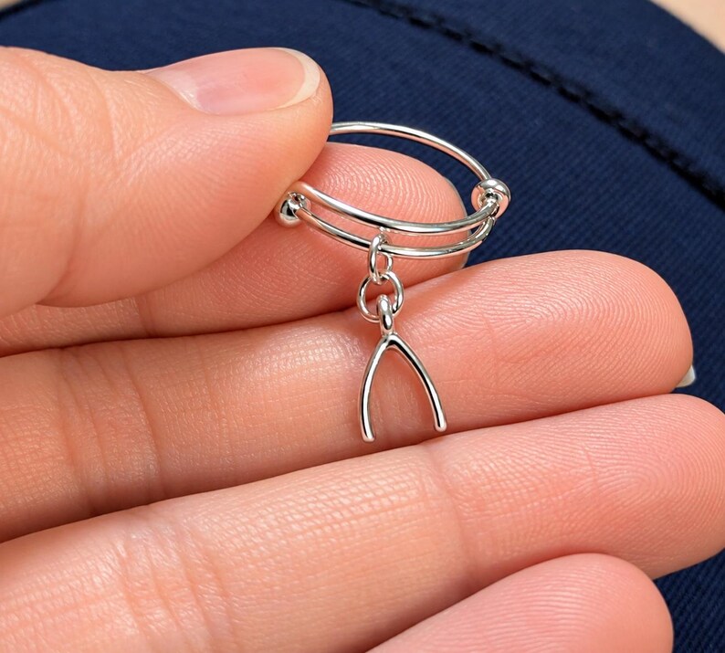 May include: A silver-toned adjustable ring featuring a delicate wishbone charm dangling from a small circular link. The band consists of two thin, parallel silver wires that overlap and terminate in small, smooth spherical beads at each end.
