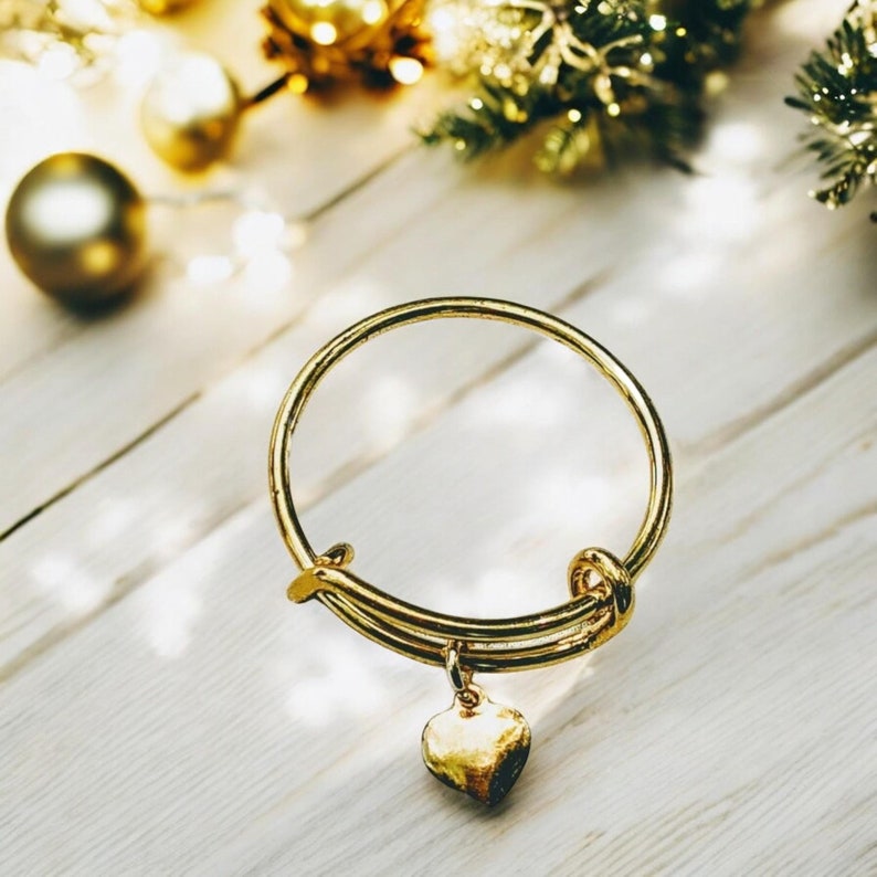 May include: A gold bangle bracelet with a heart charm. The bracelet is on a white wooden surface.