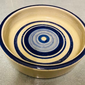 May include: A circular ceramic bowl featuring a cream-coloured interior with a dark blue spiral pattern. The rim and outer edge are also dark blue. Suitable for serving or decorative purposes.