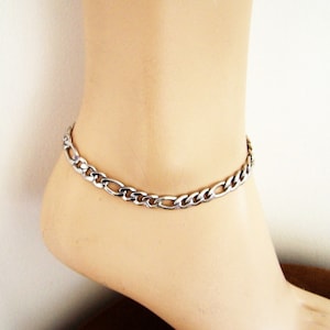 May include: A silver-colored Figaro chain anklet. The anklet is made of interlocking oval and elongated links. The jewelry is displayed on a mannequin foot against a white background.