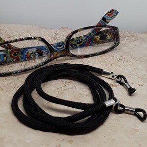 May include: Black eyeglass chain with silver clasps. The chain is made of a braided cord and is approximately 28 inches long. The chain is shown with a pair of glasses with a colorful patterned frame.