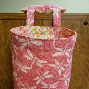 May include: A fabric storage basket with a dragonfly pattern in shades of pink and white. The interior lining features a floral pattern. The basket has a handle for hanging and is designed for organizing items.