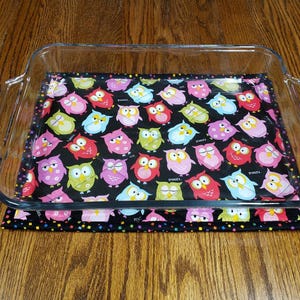 May include: A glass baking dish with a black and white owl print fabric hot pad underneath. The owls are pink, green, blue, and red with black eyes and beaks. The fabric has a confetti pattern around the border.