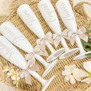 May include: Six white champagne flutes with personalised names and beige bows. The names are "Malia", "Jessica", "Kevin", "Crystal", "Lauren", and "Jenna".