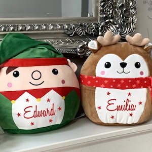 May include: Two plush Christmas-themed cushions, one shaped like an elf with a green hat and red and white trim, the other shaped like a reindeer with a red and white scarf. The elf cushion has the name "Edward" embroidered on it, and the reindeer cushion has the name "Emilia" embroidered on it.