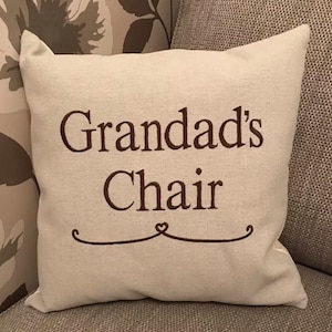 May include: A cream-coloured cushion embroidered with the words "Grandad's Chair" in brown. A decorative brown line with a heart shape is below the text. The cushion is on a grey upholstered chair.
