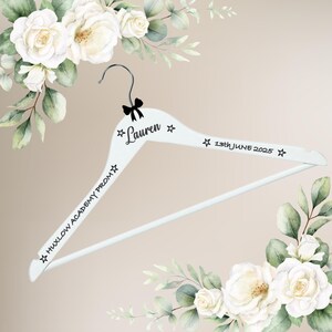 May include: White wooden clothes hanger with black text that reads "Lauren" and "13th June 2025". The hanger also has the text "Huxlow Academy Prom". A black bow is at the top of the hanger. Floral accents surround the hanger.