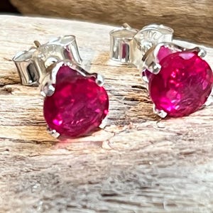 May include: A pair of stud earrings with bright, round, ruby-red gemstones set in silver metal. The earrings are presented on a textured, light brown surface, highlighting their polished look and colour. The studs are designed with a classic, elegant style.