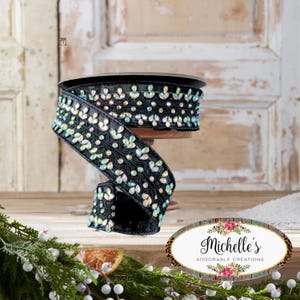 Op de afbeelding: Een rol zwart lint versierd met iriserende, juweelachtige versieringen. Het lint wordt gepresenteerd op een houten spoel. Het ontwerp heeft een herhalend patroon van bladvormige edelstenen. Een decoratief bloemen- en luipaardprintlogo staat in de rechter benedenhoek.