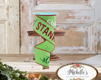 Può includere: Una bobina di nastro verde brillante con la parola "STAND" in grandi lettere rosse glitterate. Il nastro ha un bordo rosso ed è esposto su un piccolo supporto di legno. Lo sfondo include una porta rustica e vegetazione.