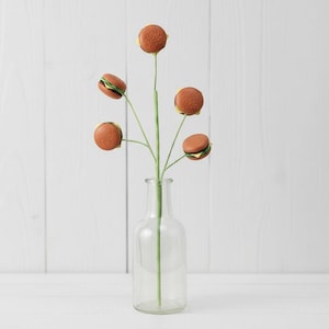 May include: A whimsical floral arrangement featuring artificial burger blooms. The 'flowers' have brown buns, green lettuce, and yellow cheese. The arrangement is in a clear glass bottle, set against a white wooden backdrop.