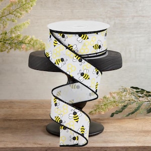 Op de afbeelding: Een rol decoratief lint met een herhalend patroon van gele bijen en honingraatvormen op een witte achtergrond. Het lint heeft een zwarte rand en wordt op een zwarte standaard gepresenteerd. Geschikt voor knutselen en cadeauverpakking.