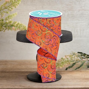 Könnte beinhalten: Eine Spule mit orangefarbenem Band mit einem schwarzen und goldenen Wirbelmuster und lila Besatz. Das Band wird auf einem schwarzen Ständer präsentiert. Die Spule hat ein türkis-weißes Etikett.