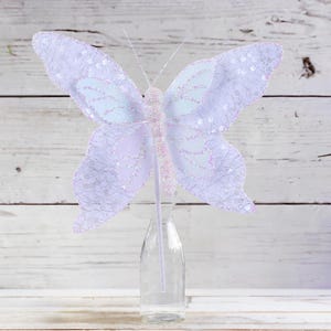 May include: A white and lavender butterfly with glitter accents is attached to a clear glass bottle. The butterfly is a decorative accent for a home or party.