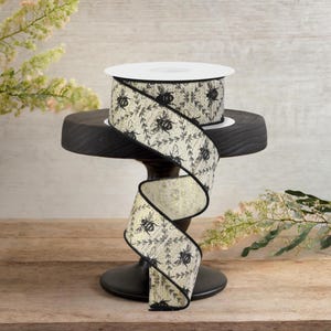 Op de afbeelding: Decoratief lint met een patroon van bijen en bladeren. Het lint is crèmekleurig met zwarte accenten en een zwarte rand. Het wordt gepresenteerd op een kleine, zwarte, ronde sokkel.