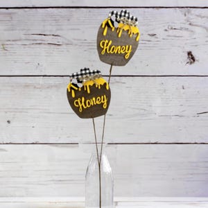 May include: Two brown honey jar shaped wooden ornaments with yellow dripping honey and black and white checkered ribbon accents. The ornaments are on brown wire stems and are in a clear glass vase.