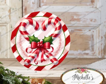 May include: A round Christmas decoration featuring two candy canes, holly, and a red bow. The border has red and white stripes. The center has a pink and white checkered pattern. The text "Michelle's aDOORable Creations" is visible.