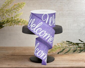 May include: A roll of purple ribbon with the word "Welcome" in white script. The ribbon is displayed on a black, round stand. The background is a light wood surface with greenery.