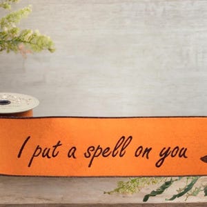 Peut inclure: Une bobine de ruban orange aux bords marron foncé, portant le texte imprimé en noir "I put a spell on you" centré entre deux illustrations de chapeaux de sorcière pointus. Le ruban est présenté horizontalement avec une extrémité enroulée.