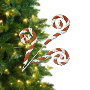 May include: Three decorative candy cane ornaments with a brown and white spiral design. The candy canes have a white, scalloped trim and are attached to a thin, silver stem. The background features a blurred Christmas tree.