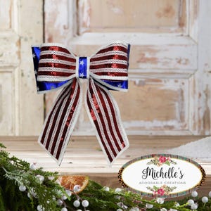 May include: A patriotic bow with red, white, and blue colors. The bow features a striped pattern with red sequins and white stripes, and blue sections with white stars. The bow is a decorative item.