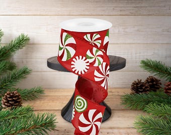 Puede incluir: Un rollo de cinta roja con un diseño de caramelos de menta blanca y verde. La cinta se muestra sobre un soporte negro, con ramas de pino y piñas en el fondo. Adecuado para decoraciones navideñas.